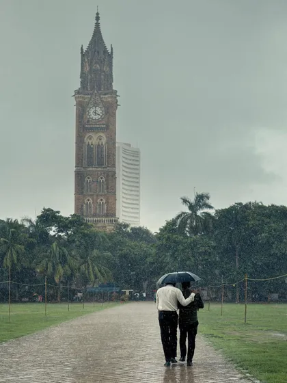 A different angle of two men walking under an umbrella at Oval Maidan, this time from further away. The composition emphasizes their smallness against the vast green field and the tall clock tower.