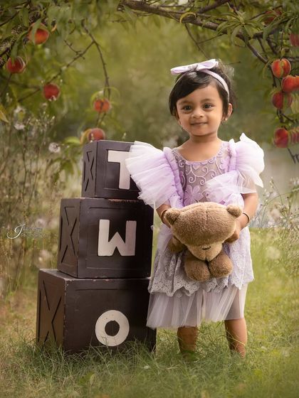 An outdoor second birthday shoot in a beautiful garden setting. The little girl in a lavender dress holds her teddy bear next to 'TWO' blocks.