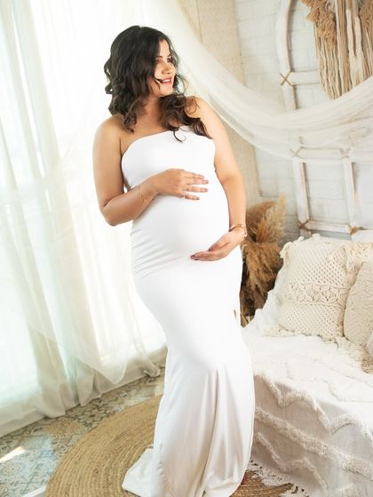 A serene solo portrait of the mom-to-be in a simple white bodycon gown. The soft, natural light from the window creates a gentle and angelic glow.