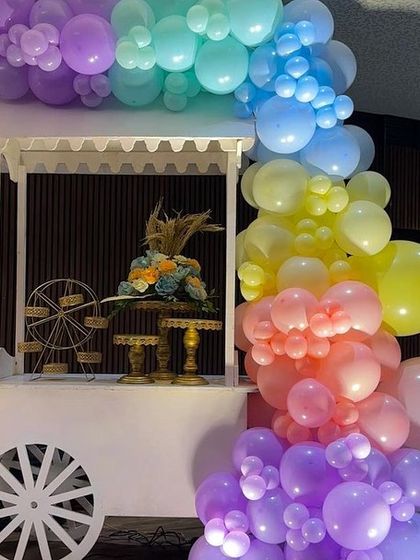The white wooden candy cart, decorated with a cascade of rainbow balloons, serves as a beautiful and functional piece of decor. It can be used to display sweets, gifts, or a cake.