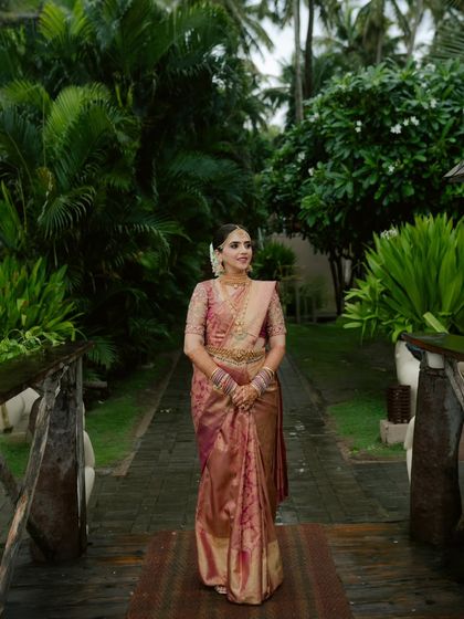 The bride walks along a path surrounded by lush tropical greenery. Her radiant smile and the rich colors of her saree are the centerpiece of this beautiful, natural portrait.