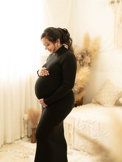 A beautiful silhouette-style portrait of an expecting mother in a black form-fitting dress. She stands in a brightly lit boho room, her profile highlighted as she lovingly holds her belly.