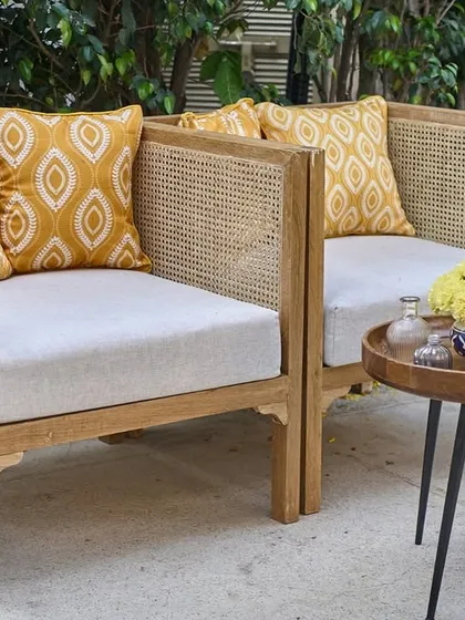 A cozy lounge corner with cane-back wooden chairs, mustard yellow patterned cushions, and a small table with a blue candle and yellow marigolds.