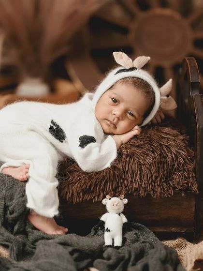 A sleepy little calf. This adorable newborn is dressed in a cow-print onesie, resting on a miniature bed in a rustic, barnyard-themed setup.