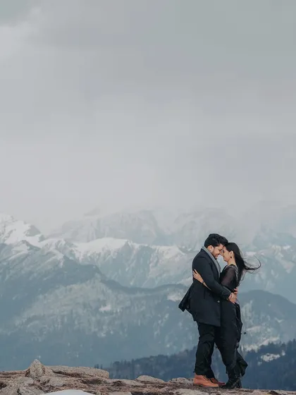 A couple embraces against a backdrop of snow-dusted mountains under a cloudy sky. The moody lighting and dramatic scenery create a powerful and romantic pre-wedding photograph.