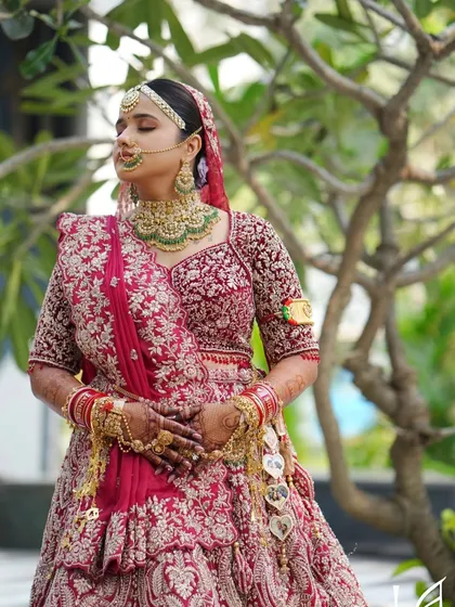 A serene outdoor portrait. The soft, natural light shows off the flawless skin work and the beautiful details of her red lehenga.