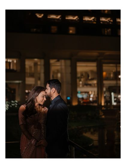 A tender moment captured at night, with the groom gently kissing the bride's forehead. The soft city lights in the background create a beautiful, romantic ambiance.