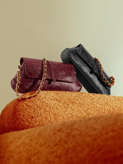 A close-up shot of two clutches with gold chain straps, styled on a textured orange surface. The composition and color contrast create a warm, luxurious, and on-trend image.