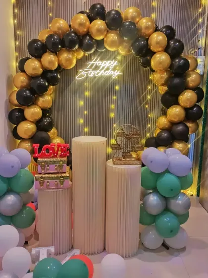 A classic and elegant birthday decoration with a black and gold balloon arch. The setup is completed with plinth stands, decorative props, and smaller balloon clusters.