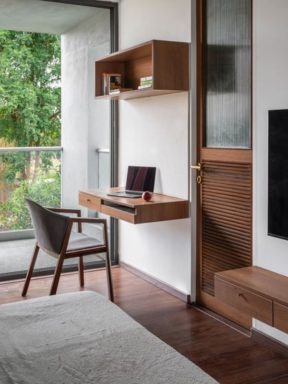 A compact work-from-home nook designed within a bedroom, featuring a floating teak desk and shelf. The space opens onto a balcony, providing natural light and a connection to the outdoors.