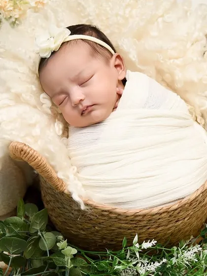A close-up portrait of a baby swaddled in white, sleeping in a basket surrounded by white pumpkins and greenery. The focus is on the baby's serene expression.