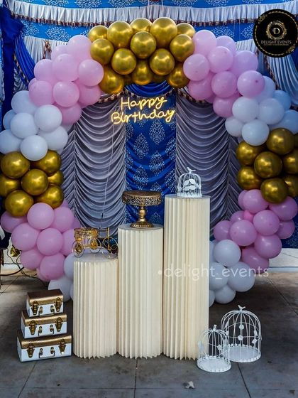 A classic and regal color combination of pink, white, and gold balloons set against a royal blue and white draped backdrop. This circular arch setup includes a neon sign and elegant pedestals, perfect for a milestone celebration.