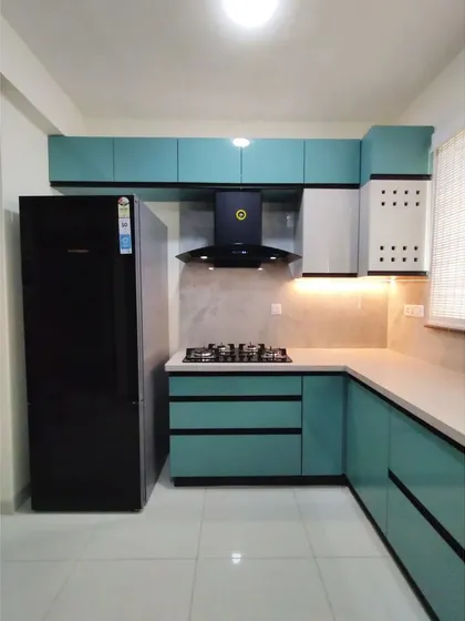 A kitchen should be as bright and beautiful as the life it sustains. This bespoke kitchen in a vibrant teal features a highly functional L-shaped layout, complete with a chimney, four-burner hob, and ample overhead and base cabinets, proving that practicality and personality can go hand in hand.