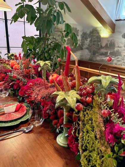 A full view of the long dining table, showcasing the impressive scale of the central floral and fruit installation. The design brings a sense of natural, untamed opulence indoors.