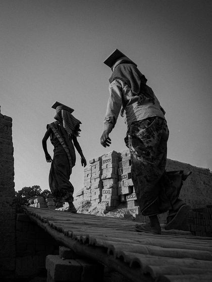 Two women walk along a rampart of bricks, their forms creating strong silhouettes against the sky. The low angle emphasizes their journey and the scale of the structures they build.