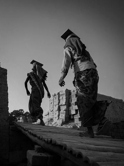 Two women walk along a rampart of bricks, their forms creating strong silhouettes against the sky. The low angle emphasizes their journey and the scale of the structures they build.