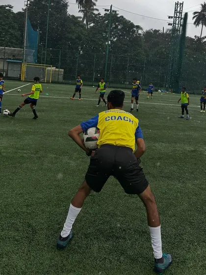Storms don't stop us, they build our character. A coach stands ready with a ball as our youth players continue their training session with intensity, even in the pouring rain, developing the resilience required to succeed.