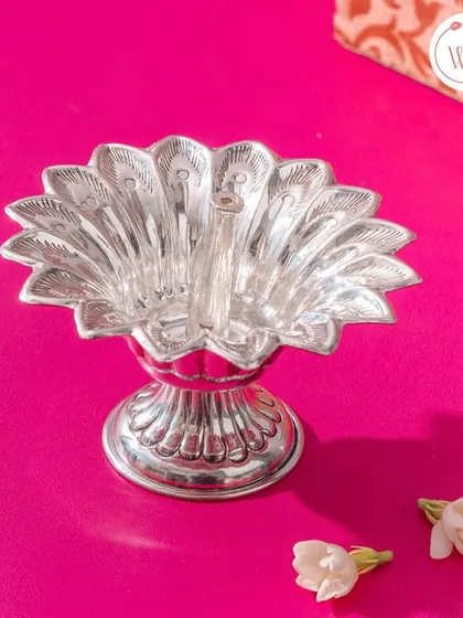A pure silver peacock-shaped diya (oil lamp), showcasing intricate feather details. It's a beautiful item for a puja room or as a traditional gift.