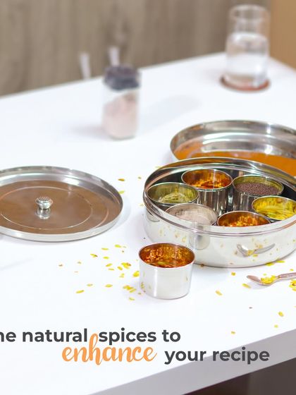 A lifestyle shot of a stainless steel masala dabba (spice box) on a clean white kitchen table. This image shows the product's function and capacity in a realistic setting.