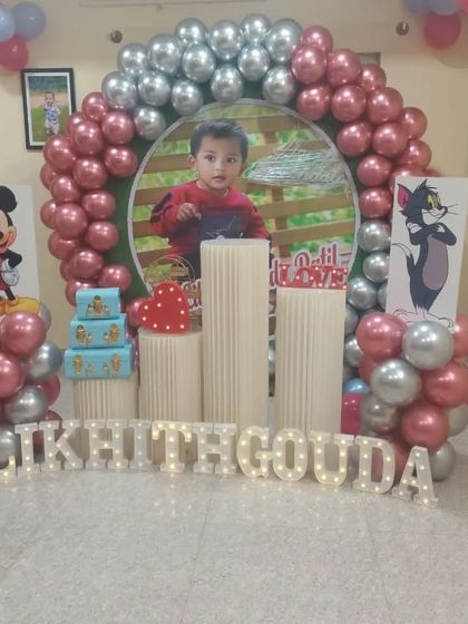 A first birthday party setup featuring a personalized photo backdrop of the birthday boy. The decor includes Mickey Mouse and Tom & Jerry cutouts and light-up letters.