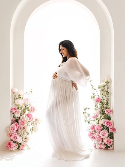 A side profile of the mom-to-be in the floral archway, cradling her bump. The combination of the white gown and pink flowers is simply perfect.
