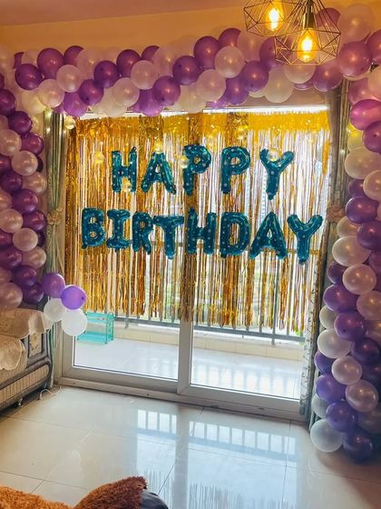A purple and white balloon arch set up against a balcony door. This decoration uses a gold tinsel curtain and blue 'Happy Birthday' foil letters for contrast.
