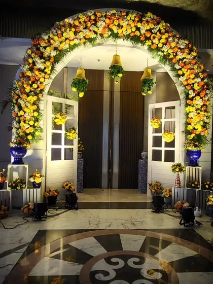 An elegant Haldi entrance combining traditional and modern elements. A lush floral arch made of marigolds is paired with white doors and blue pottery props, creating a fresh and stylish welcome.