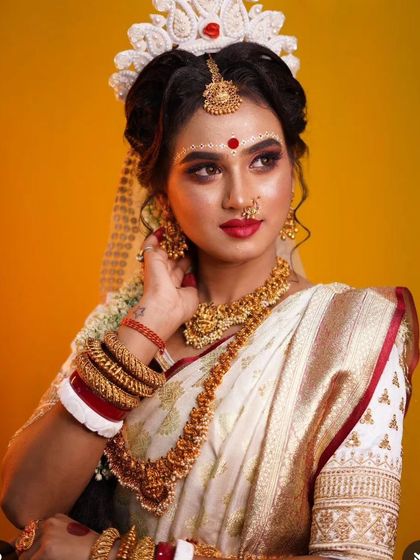 A beautiful Bengali bride in a white and red saree, wearing traditional gold-plated jewellery. The look is defined by the bindi design and the 'mukut'.