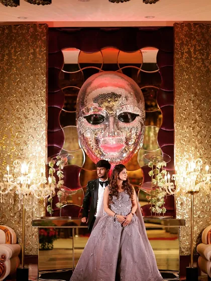 A grand portrait of a couple at their reception, posing in front of a large, artistic mask decoration.