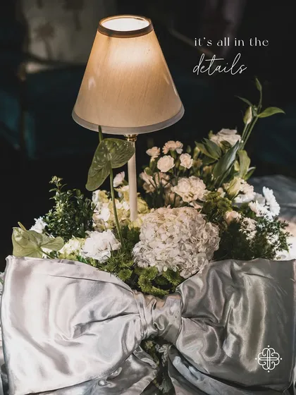 A detail of the elegant decor from a seaside soirée, with a table lamp nestled within a white and green floral arrangement and a large silver bow.