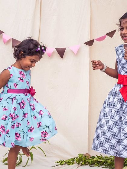 A playful moment between two girls in their custom dresses. One wears a light blue floral, while the other is in a black-and-white check with a bold red bow, showcasing their individual styles.