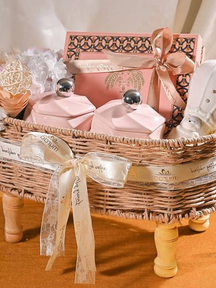 This elegant baby announcement hamper uses a soft, neutral color palette. It features a wicker basket filled with pink ceramic jars, a hot-air balloon diffuser, and is tied with a beautiful satin and lace ribbon.