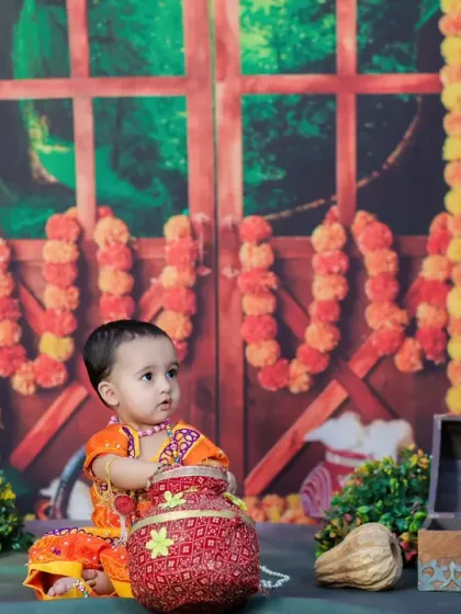 A baby dressed in a traditional orange outfit for a Janmashtami shoot, looking thoughtfully to the side while holding a decorated pot.