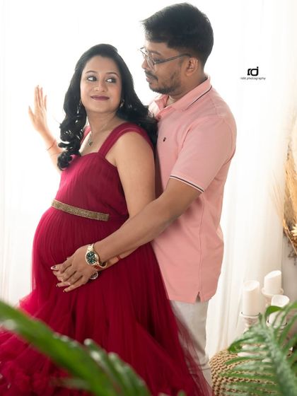 A classic couple's pose by the window. The soft, diffused light and the rich red of the gown create a warm and romantic atmosphere for this pregnancy portrait.