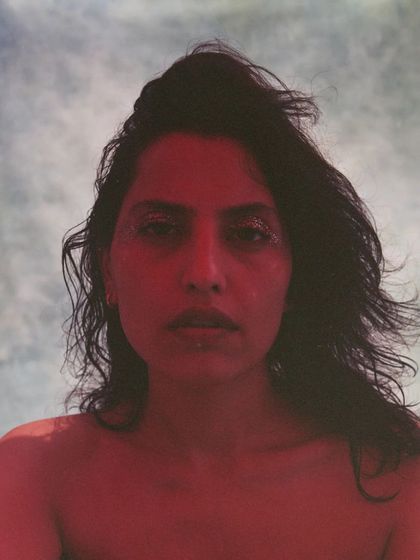 A raw and intense portrait from an editorial shoot. The styling is minimal, focusing on the model's expression and the dramatic red lighting, with a touch of glitter on the eyes to catch the light.