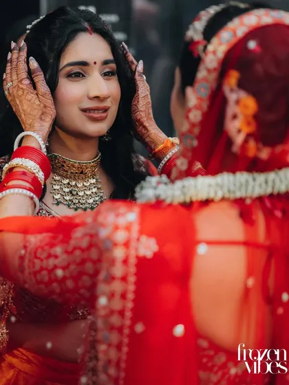 A bride looks at her reflection in a mirror held by her mother, who is dressed in a traditional red saree. This is a poignant, symbolic moment of final preparations and maternal love.