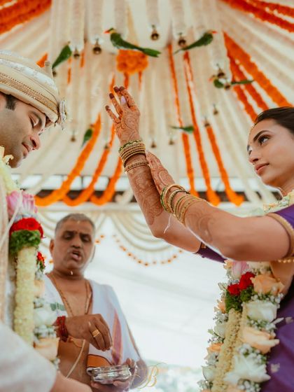 A candid moment during the wedding ceremony, with the mandap's hanging garlands of marigold, tuberose, and leaf parrots creating a beautiful overhead texture.