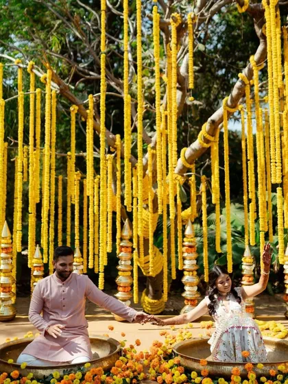 A fun and festive shot of the couple sitting in traditional brass pots during their Haldi ceremony, surrounded by marigold decorations. This captures the unique and playful elements of the celebration.