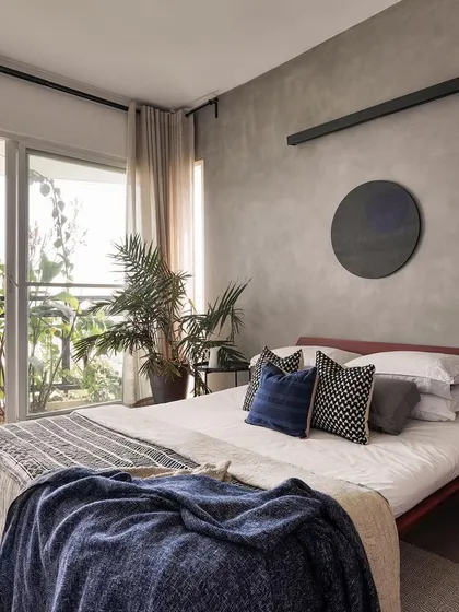 A tranquil bedroom from the 'Rustic Minimal' apartment, where a cement-textured wall contrasts with warm wood and soft textiles. The full-height glass door opens onto a lush private balcony, blurring the line between inside and out. Our focus here was on creating a personal sanctuary that feels both minimalist and deeply comfortable.