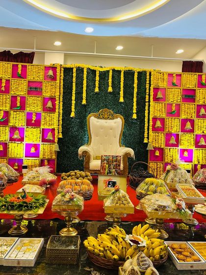 This vibrant Seemantham stage features a checkered backdrop in yellow and magenta, with hanging marigold garlands and brass bells. The foreground is filled with beautifully arranged traditional offerings.