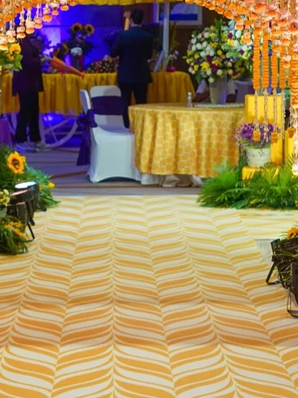 The entrance walkway for the Haldi ceremony, featuring a yellow and white patterned carpet runner. The path is lined with sunflowers and illuminated by spotlights.
