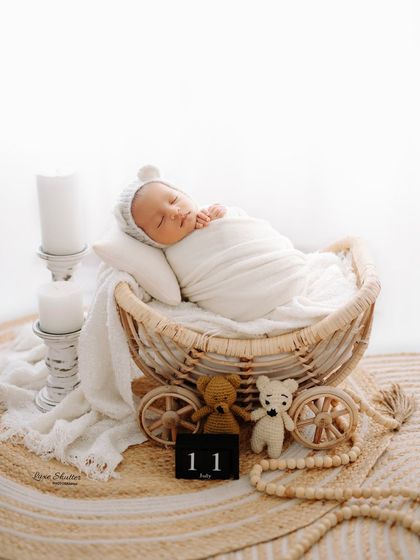An all-white, serene newborn setup. The baby is swaddled and sleeping in a rustic wheeled basket, accompanied by teddy bears and a date block marking his arrival.