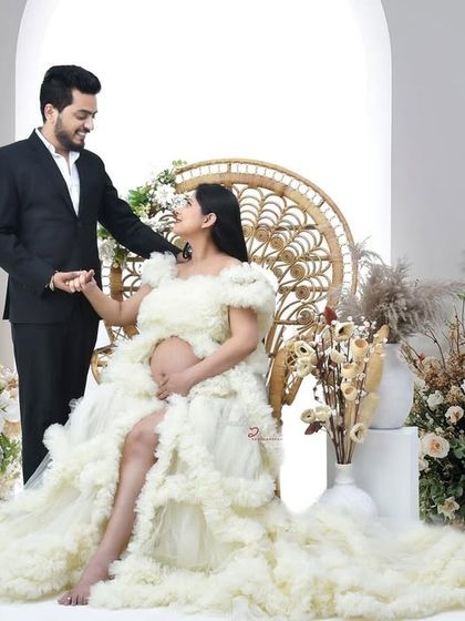 A romantic couple's portrait from a maternity session. The mother-to-be is seated in a beautiful white gown while her partner stands beside her, creating a classic and loving image.