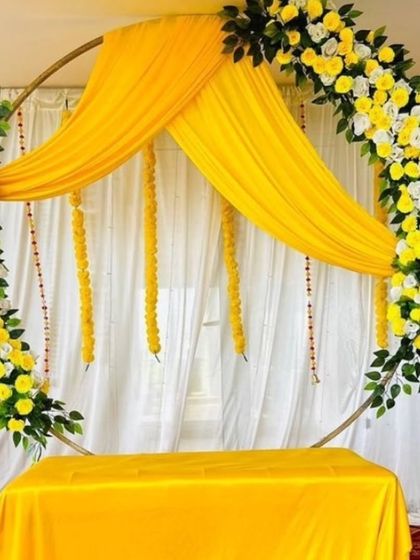 A simple and elegant ring backdrop for a Haldi or engagement ceremony. The frame is decorated with yellow and white flowers and draped with yellow fabric for a soft, romantic feel.