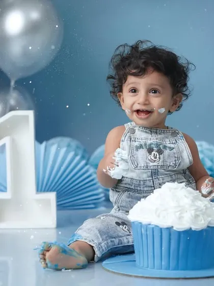A baby boy in Mickey Mouse overalls has a blast smashing a giant blue cupcake against a festive blue and silver balloon backdrop.