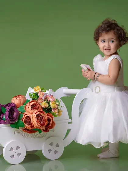 A classic portrait of a little girl in a white dress with a flower-filled pram. The solid green background provides a beautiful contrast and a touch of nature.