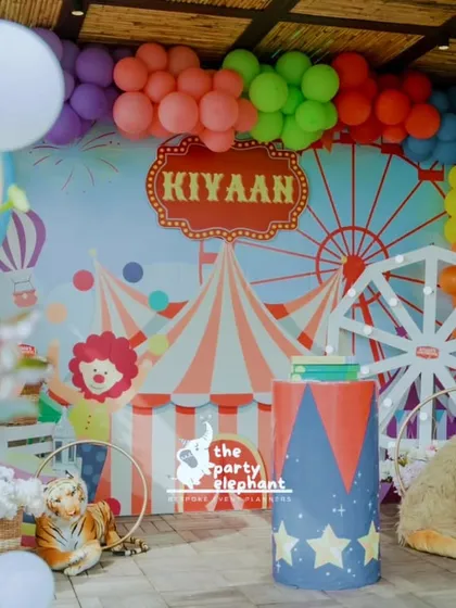 Step right up to a whimsical carnival birthday. This vibrant backdrop features a custom circus tent and ferris wheel design, framed by a colorful balloon garland and playful animal props, setting the stage for a day of fun and games.