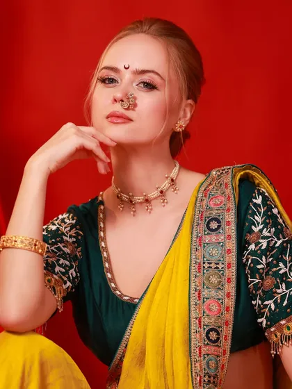 A close-up beauty shot of the model in a festive saree, highlighting her traditional nose ring and jewelry against a bold red backdrop.