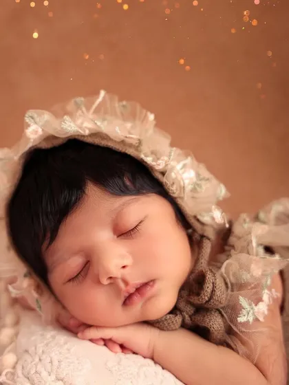 A close-up portrait of a sleeping newborn wearing an elegant, vintage-style lace bonnet. The soft focus and warm tones give this image a timeless, heirloom quality.