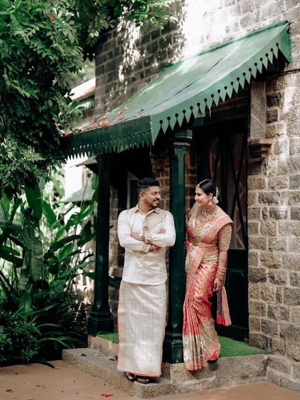 A couple posing outside a traditional stone house. His simple white dhoti-kurta and her peach saree create a look of timeless elegance.